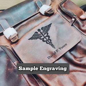 May include: A brown leather backpack with a zippered pocket on the front. The pocket has a black engraved medical symbol and the text "David P. Vance".