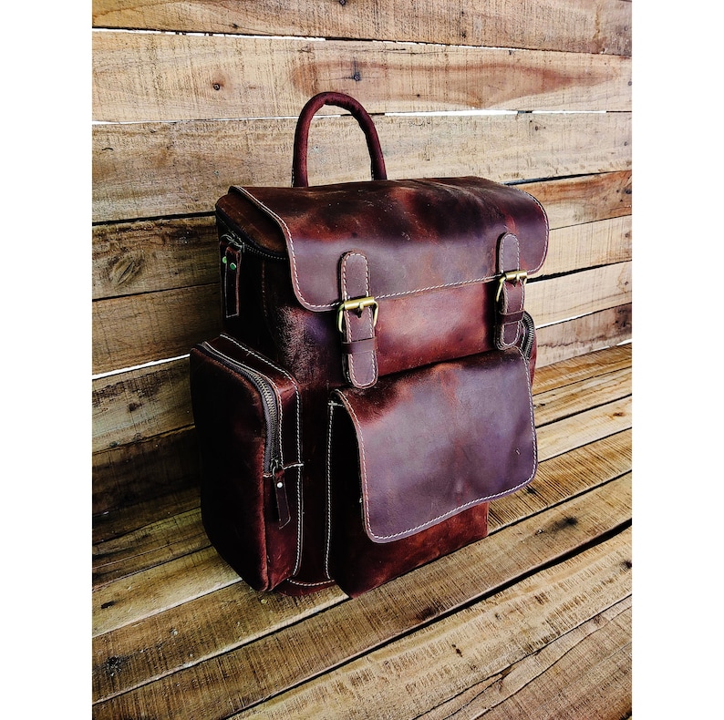 May include: A brown leather backpack with two large front pockets and two adjustable buckles. The backpack has a top handle and a zipper closure.