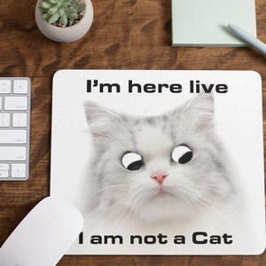 May include: A white mouse mat featuring a white cat with large, black eyes. The cat is looking at the camera with a serious expression. The text "I'm here live I am not a Cat" is printed on the mouse mat.
