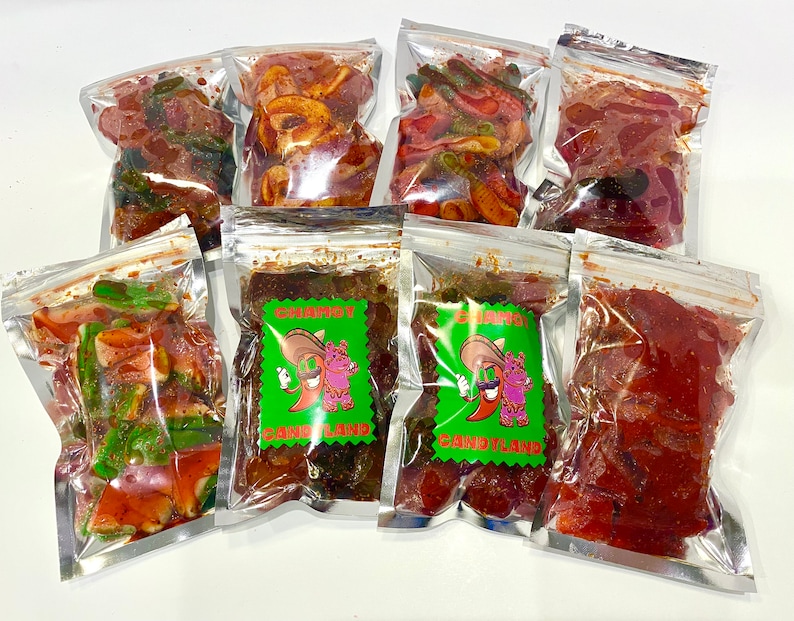 May include: Seven clear plastic bags filled with different types of sweets. Some bags contain green and red sweets, while others have brown and red sweets. Two bags have a green label with a cartoon chili pepper wearing a sombrero and the text "Gummy Candyland."
