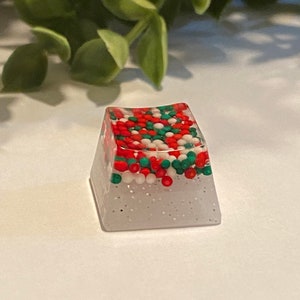 May include: A clear, square-shaped keyboard keycap filled with red, green, and white sprinkles. The keycap has a glittery, translucent appearance. The background features green foliage and a white surface.