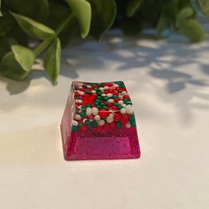 May include: A rectangular, clear resin keycap with a pink glitter base and a top layer of red, green, and white spherical sprinkles. The keycap is a custom design, likely for a mechanical keyboard.