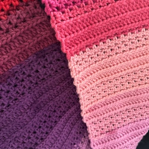 May include: A hand-crocheted textile featuring a vibrant gradient of colors transitioning from deep purple and burgundy to bright pink, coral, and light peach. The fabric displays a consistent ribbed texture created by repeating crochet stitches.