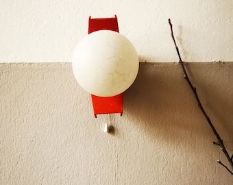 Op de afbeelding: Een rode wandlamp met een witte bolvormige lamp. De lamp heeft een eenvoudig ontwerp en is bevestigd aan een witte muur.