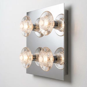 May include: A square wall-mounted light fixture featuring a reflective chrome backplate and four illuminated clear glass knobs arranged in two rows. Each knob has a textured, circular pattern and is mounted on a metallic base, emitting a warm glow.