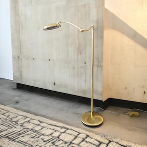 Può includere: Una lampada da terra in ottone con un braccio curvo e un paralume circolare. La lampada ha una base rotonda e un palo sottile. La lampada è posizionata davanti a un muro di cemento e un tappeto fantasia.