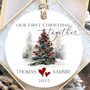 May include: A white Christmas ornament with the text "OUR FIRST CHRISTMAS TOGETHER". It features a watercolor painting of a Christmas tree with red ornaments and two snow-covered trees. The names "THOMAS" and "SAMMIE" are below the tree, along with two hearts and the year "2025".