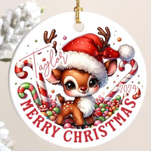 May include: A round Christmas ornament featuring a cartoon reindeer wearing a Santa hat and surrounded by candy canes and colorful candies. The ornament has the text "Merry Christmas" and the name "Taylor" on it, with the year "2024".