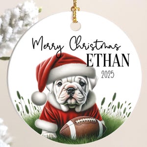 May include: A round white Christmas ornament featuring a bulldog wearing a Santa hat and coat, holding a football. The ornament has the text "Merry Christmas ETHAN 2025" in black script.