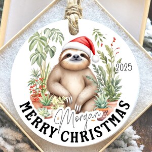 May include: A round Christmas ornament featuring a smiling sloth wearing a Santa hat, surrounded by potted plants. The ornament has the text "Merry Christmas Morgan 2025" in black and white.