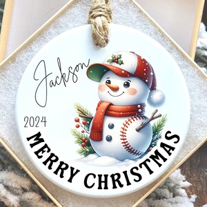 May include: A white ceramic Christmas ornament featuring a snowman wearing a baseball cap and a red scarf. The ornament has the name "Jackson" and the year "2024" printed on it, with "MERRY CHRISTMAS" in a curved font.