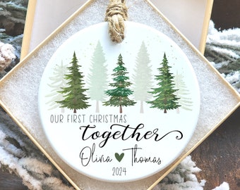 May include: A white ceramic Christmas ornament with a forest design and the text "Our First Christmas Together". The ornament includes the names Olivia and Thomas, a green heart, and the year 2024. It hangs from a jute cord and is in a gift box.