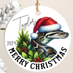 May include: A white ceramic Christmas ornament with a snake wearing a Santa hat. The ornament has the text "Taylor" and "2024 Merry Christmas" on it.