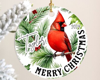 May include: A round Christmas ornament featuring a vibrant red cardinal perched on a branch, surrounded by green pine needles and holly. The ornament includes the text "Merry Christmas" and "2024", with the name "Taylor" in a script font. A gold string is attached for hanging.