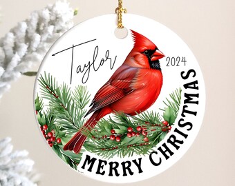 May include: A white ceramic Christmas ornament featuring a vibrant red cardinal perched on a pine branch with red berries. The ornament has the text "Merry Christmas" and "2024" with the name "Taylor" in script.