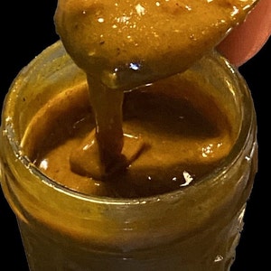 May include: A close-up of a glass jar filled with a thick, golden brown sauce. The sauce is being scooped out with a spoon, revealing its smooth and creamy texture.