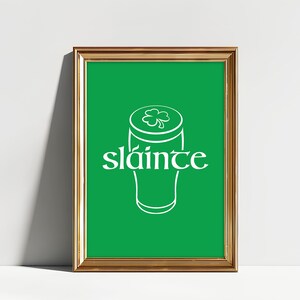 K&ouml;nnte beinhalten: Ein gerahmter Druck mit einem goldfarbenen Rahmen. Der Druck zeigt einen gr&uuml;nen Hintergrund mit einer wei&szlig;en Umrisszeichnung eines Bierkrugs und dem Wort "sl&aacute;inte" in Wei&szlig;, ein g&auml;lisches Wort f&uuml;r "Prost". Ein Kleeblatt befindet sich oben auf dem Bierkrug.