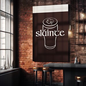 K&ouml;nnte beinhalten: Ein dunkelbraunes Schild mit dem Wort "sl&aacute;inte" in wei&szlig;er Schrift, &uuml;ber einer wei&szlig;en Umrisszeichnung eines Bierglases mit einem Kleeblatt. Das Schild h&auml;ngt an einer Backsteinmauer in einer Bar.