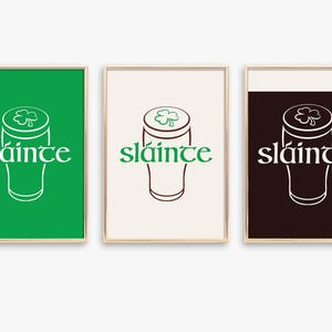 K&ouml;nnte beinhalten: Drei gerahmte Kunstdrucke mit goldenen R&auml;ndern. Jeder Druck zeigt ein stilisiertes Pintglas mit einem Kleeblatt und dem Wort "sl&aacute;inte" in einer dekorativen Schriftart. Die Hintergr&uuml;nde sind gr&uuml;n, cremefarben und dunkelbraun.