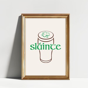 K&ouml;nnte beinhalten: Gerahmter Druck mit goldenem Rahmen. Der Druck zeigt eine braune Umrisszeichnung eines Bierkrugs, gekr&ouml;nt von einem gr&uuml;nen Kleeblatt. Das Wort "sl&aacute;inte" steht in gr&uuml;ner Schrift darunter.