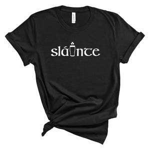 Puede incluir: Camiseta negra con la palabra "sl&aacute;inte" en blanco, que significa "salud" en ga&eacute;lico. El dise&ntilde;o incluye un tr&eacute;bol y un vaso de pinta. La camiseta est&aacute; hecha de un material suave.