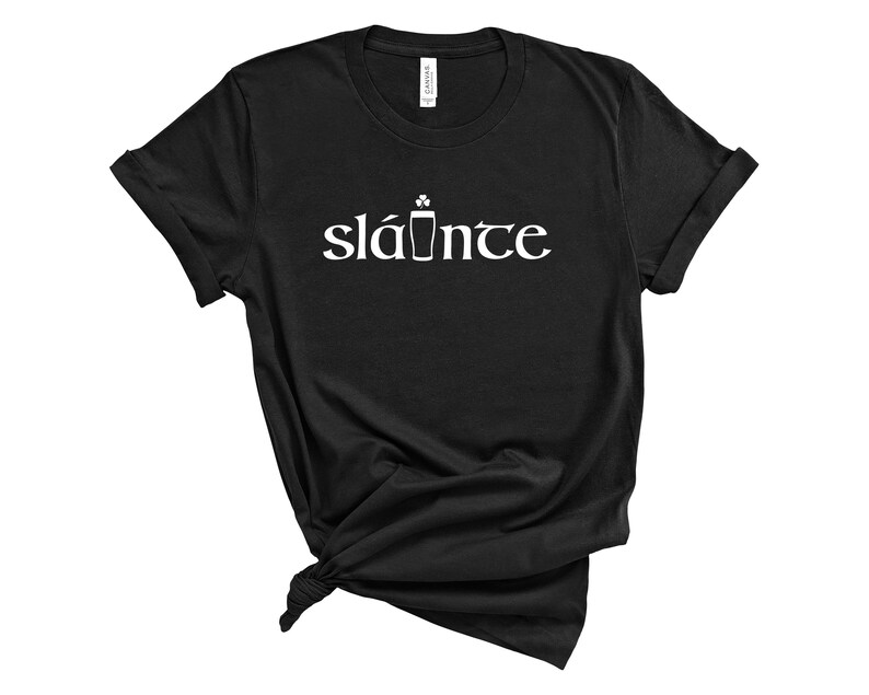 Puede incluir: Camiseta negra con la palabra "sl&aacute;inte" en blanco, que significa "salud" en ga&eacute;lico. El dise&ntilde;o incluye un tr&eacute;bol y un vaso de pinta. La camiseta est&aacute; hecha de un material suave.