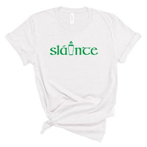 Puede incluir: Camiseta blanca con la palabra "sl&aacute;inte" en verde, un brindis tradicional irland&eacute;s en ga&eacute;lico. La palabra est&aacute; estilizada con un tr&eacute;bol y un s&iacute;mbolo de pinta. La camiseta es de un material suave.
