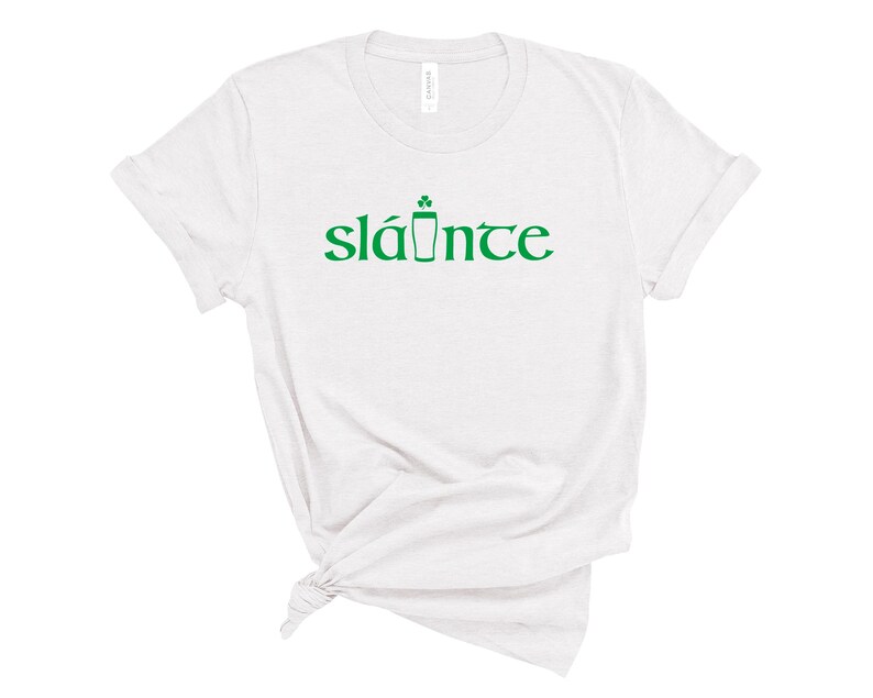 Puede incluir: Camiseta blanca con la palabra "sl&aacute;inte" en verde, un brindis tradicional irland&eacute;s en ga&eacute;lico. La palabra est&aacute; estilizada con un tr&eacute;bol y un s&iacute;mbolo de pinta. La camiseta es de un material suave.