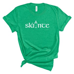 Puede incluir: Camiseta verde con la palabra "sl&aacute;inte" en blanco, un brindis tradicional irland&eacute;s en ga&eacute;lico. La camiseta tiene cuello redondo y mangas enrolladas. La palabra "sl&aacute;inte" est&aacute; escrita en una fuente estilizada con un tr&eacute;bol y un s&iacute;mbolo de pinta.