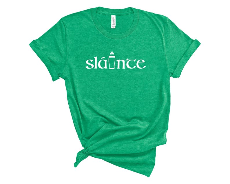 Puede incluir: Camiseta verde con la palabra "sl&aacute;inte" en blanco, un brindis tradicional irland&eacute;s en ga&eacute;lico. La camiseta tiene cuello redondo y mangas enrolladas. La palabra "sl&aacute;inte" est&aacute; escrita en una fuente estilizada con un tr&eacute;bol y un s&iacute;mbolo de pinta.