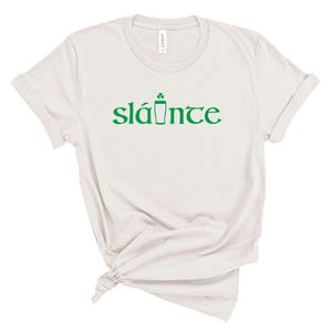 Puede incluir: Una camiseta de color claro con la palabra "sl&aacute;inte" en verde, un brindis tradicional irland&eacute;s. La palabra va acompa&ntilde;ada de un tr&eacute;bol verde y un dibujo de pinta. La camiseta est&aacute; hecha de un material suave.