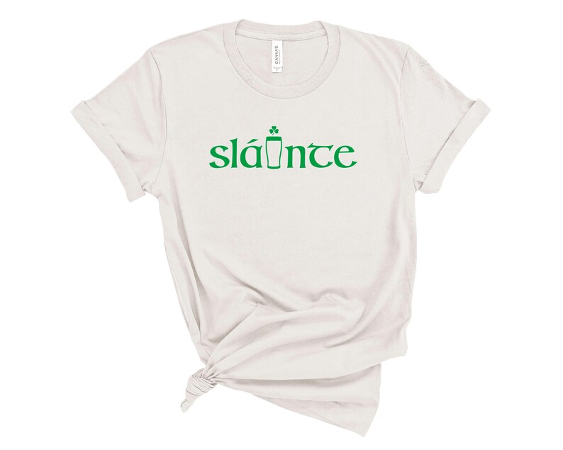 Puede incluir: Una camiseta de color claro con la palabra "sl&aacute;inte" en verde, un brindis tradicional irland&eacute;s. La palabra va acompa&ntilde;ada de un tr&eacute;bol verde y un dibujo de pinta. La camiseta est&aacute; hecha de un material suave.