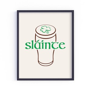 K&ouml;nnte beinhalten: Gerahmter Kunstdruck mit einer braunen Umrisszeichnung eines Bierkrugs und einem gr&uuml;nen Kleeblatt. Das Wort "sl&aacute;inte" steht in gr&uuml;ner Schrift darunter. Der Hintergrund ist hellbeige.
