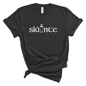 Puede incluir: Camiseta gris oscuro con la palabra "sl&aacute;inte" en blanco, una palabra ga&eacute;lica que significa "salud". La palabra est&aacute; estilizada con un tr&eacute;bol y un s&iacute;mbolo de pinta. La camiseta es de un material suave y tiene las mangas enrolladas.