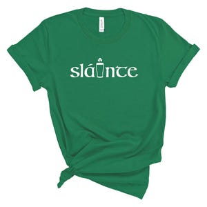 Puede incluir: Una camiseta verde con la palabra "sl&aacute;inte" en blanco, con un dise&ntilde;o de tr&eacute;bol y pinta de cerveza. La camiseta es de material suave y tiene las mangas enrolladas. Una camiseta informal para el D&iacute;a de San Patricio.