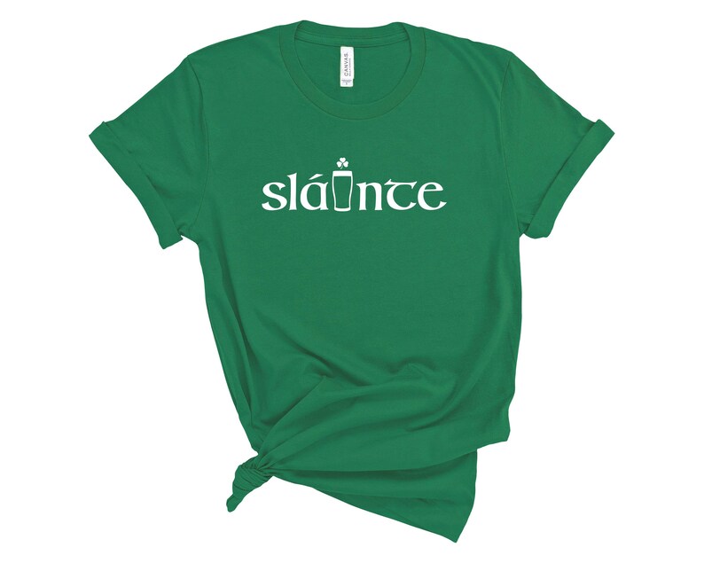 Puede incluir: Una camiseta verde con la palabra "sl&aacute;inte" en blanco, con un dise&ntilde;o de tr&eacute;bol y pinta de cerveza. La camiseta es de material suave y tiene las mangas enrolladas. Una camiseta informal para el D&iacute;a de San Patricio.