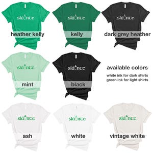 Puede incluir: Una colecci&oacute;n de camisetas en varios colores, incluyendo verde, negro y blanco. Cada camiseta presenta la palabra "sl&aacute;inte" en una fuente estilizada. Los colores disponibles se enumeran con tinta blanca para camisas oscuras y tinta verde para camisas claras.