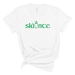 Puede incluir: Camiseta blanca con la palabra "sl&aacute;inte" en verde, con un dise&ntilde;o de tr&eacute;bol y pinta de cerveza. La camiseta est&aacute; hecha de un material suave y tiene un cuello redondo cl&aacute;sico.