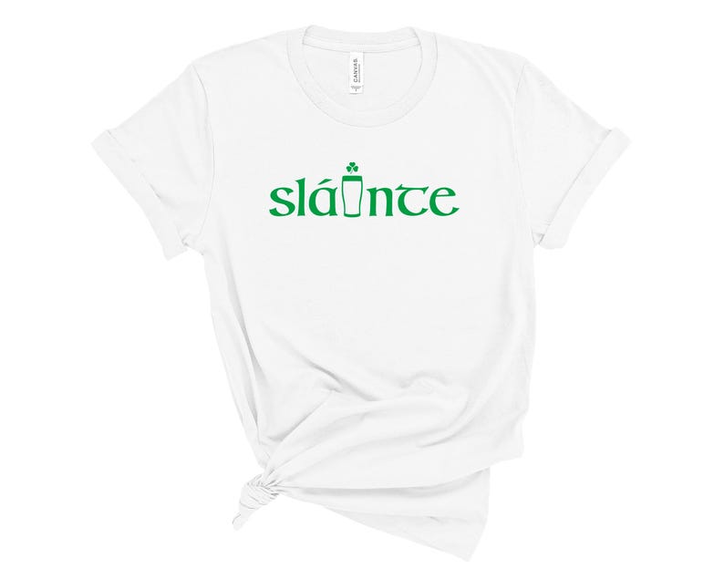 Puede incluir: Camiseta blanca con la palabra "sl&aacute;inte" en verde, con un dise&ntilde;o de tr&eacute;bol y pinta de cerveza. La camiseta est&aacute; hecha de un material suave y tiene un cuello redondo cl&aacute;sico.