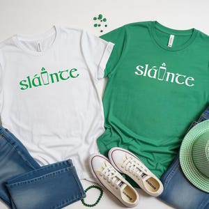 Puede incluir: Dos camisetas, una blanca y otra verde, con la palabra "sl&aacute;inte" en verde y blanco, respectivamente. La palabra va acompa&ntilde;ada de un gr&aacute;fico de una pinta y un tr&eacute;bol. Las camisetas se combinan con vaqueros, zapatos y un sombrero.
