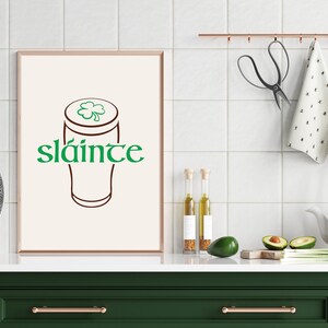 K&ouml;nnte beinhalten: Ein gerahmter Druck mit einem Bierkrug-Design und dem Wort "sl&aacute;inte" in Gr&uuml;n. Das Kunstwerk wird in einer K&uuml;chenumgebung mit einer gr&uuml;nen Arbeitsplatte und einer wei&szlig;en Fliesenspiegel pr&auml;sentiert. Weitere K&uuml;chenartikel sind sichtbar.