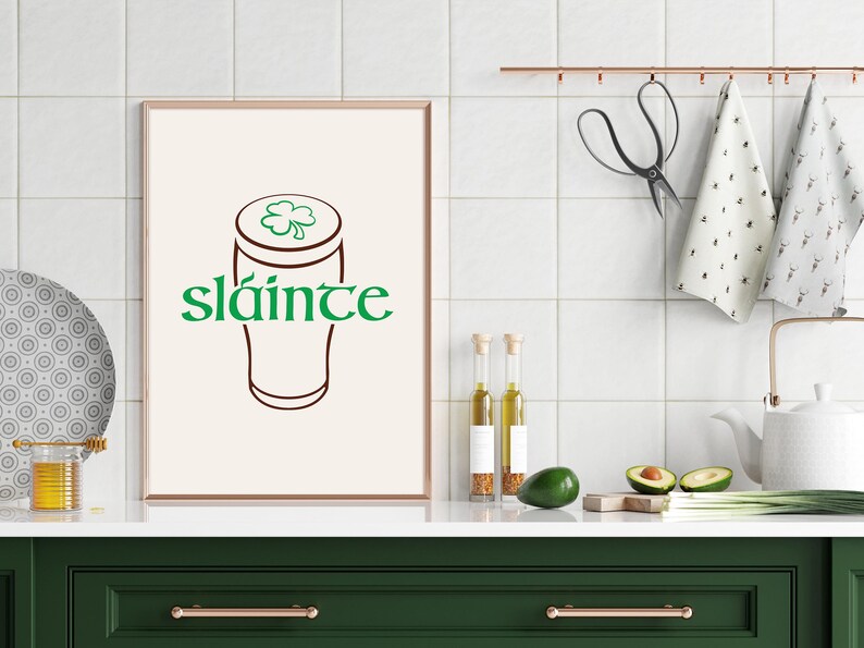 K&ouml;nnte beinhalten: Ein gerahmter Druck mit einem Bierkrug-Design und dem Wort "sl&aacute;inte" in Gr&uuml;n. Das Kunstwerk wird in einer K&uuml;chenumgebung mit einer gr&uuml;nen Arbeitsplatte und einer wei&szlig;en Fliesenspiegel pr&auml;sentiert. Weitere K&uuml;chenartikel sind sichtbar.