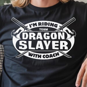 May include: Black t-shirt with a white graphic design. The design features the text "I'M RIDING TEAM DRAGON SLAYER WITH COACH" in a stylized font, along with two crossed swords and a dragon head.