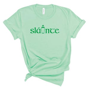 Puede incluir: Una camiseta verde claro con la palabra "sl&aacute;inte" impresa en verde. La palabra est&aacute; estilizada con un tr&eacute;bol y un icono de pinta. La camiseta est&aacute; hecha de un material suave y tiene mangas enrolladas.