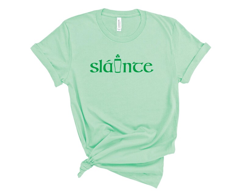 Puede incluir: Una camiseta verde claro con la palabra "sl&aacute;inte" impresa en verde. La palabra est&aacute; estilizada con un tr&eacute;bol y un icono de pinta. La camiseta est&aacute; hecha de un material suave y tiene mangas enrolladas.