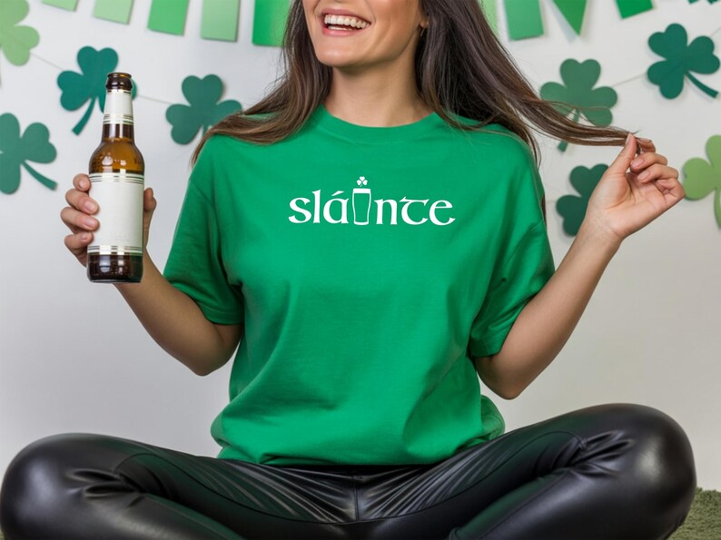 Puede incluir: Una camiseta verde con la palabra "sl&aacute;inte" en blanco, con un icono de vaso de cerveza. La persona sostiene una botella de cerveza con una etiqueta en blanco. El fondo incluye decoraciones de tr&eacute;boles, perfecto para el D&iacute;a de San Patricio.