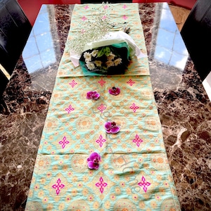 May include: A light blue table runner with a gold and pink geometric pattern. The runner is decorated with a bouquet of white flowers and pink orchid blossoms.