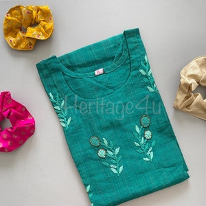 May include: A teal blue cotton top with embroidered green leaves and white beads. The top has a round neckline and is pictured with three hair scrunchies in pink, gold, and yellow.