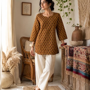 May include: Mustard-colored tunic with a red floral pattern and three-quarter sleeves. Paired with cream-colored pants and brown sandals. The outfit is worn in a well-lit room with natural decor.