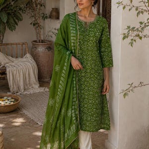 May include: A green cotton kurta set with a matching dupatta. The kurta features a white floral pattern and three-quarter sleeves. The set is paired with white pants. The model is wearing brown sandals.